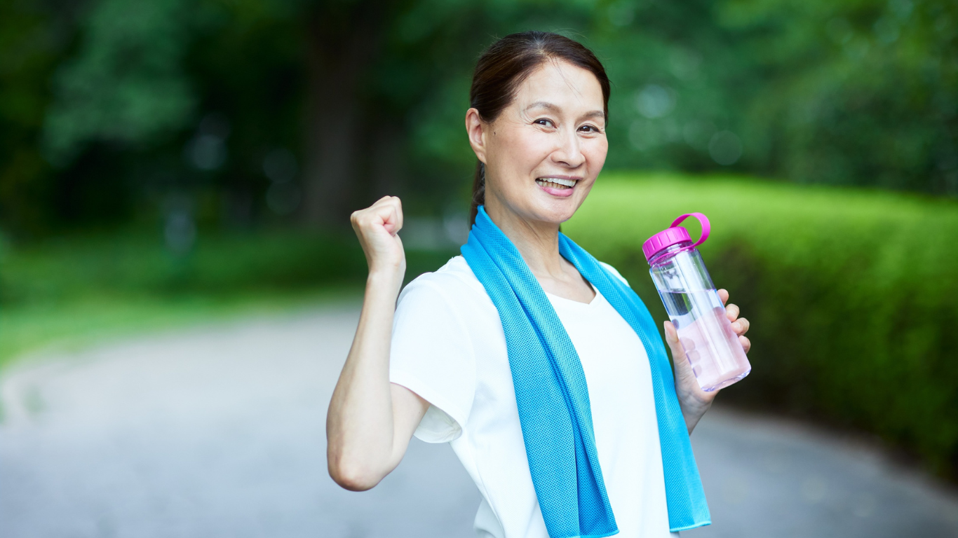 Une femme souriante en tenue de sport, tenant une bouteille d'eau, avec une serviette bleue autour du cou, dans un parc.