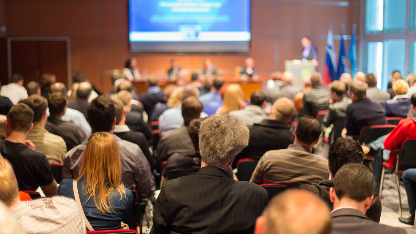 Une salle de conférence remplie de participants assis face à une scène avec un panneau d'affichage et des drapeaux à l'arrière, une personne parle au micro.