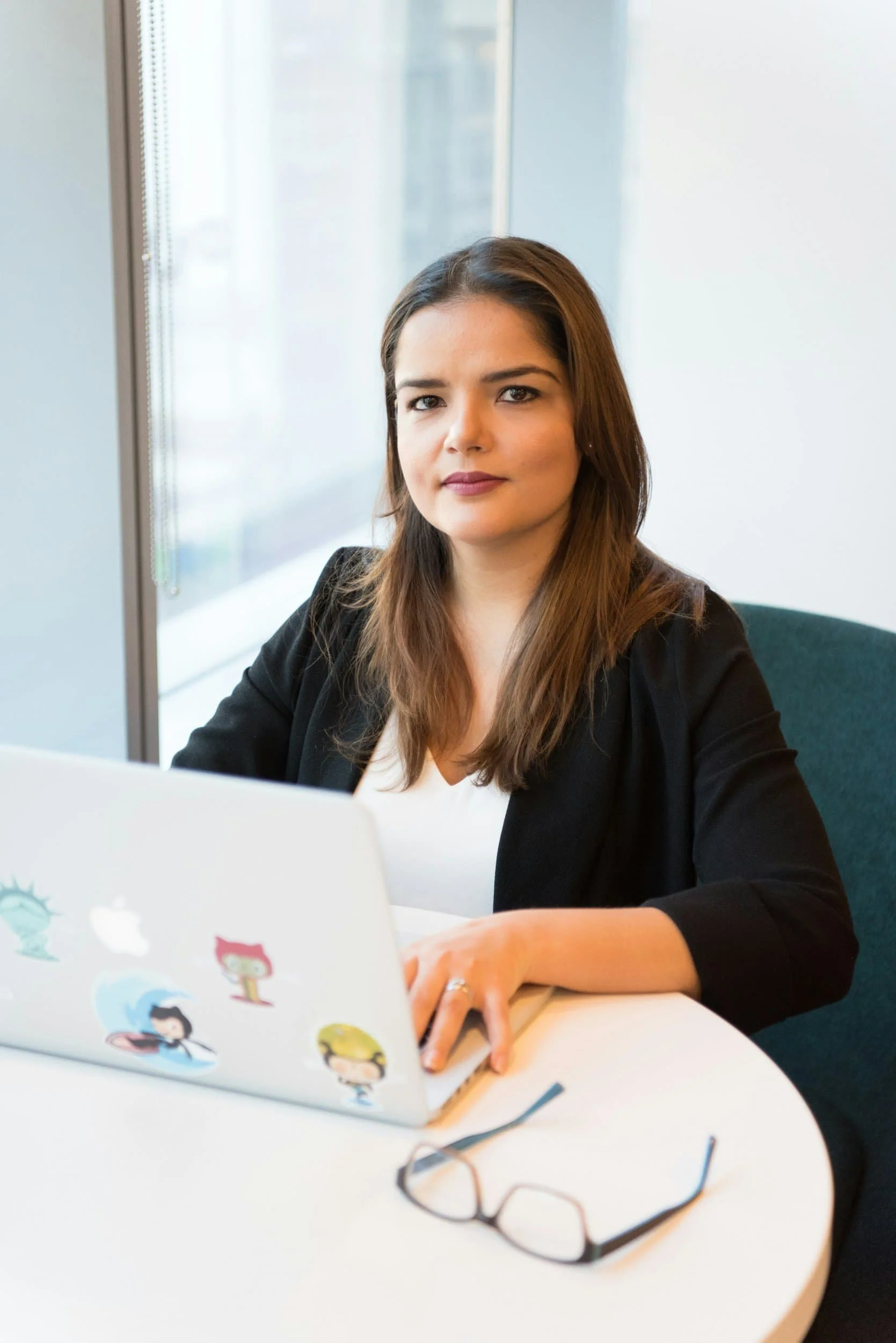 A woman with long brown hair working at a white table with a silver laptop decorated with colorful cartoon character stickers, a pair of glasses, and a pen, in a modern office with large windows.
