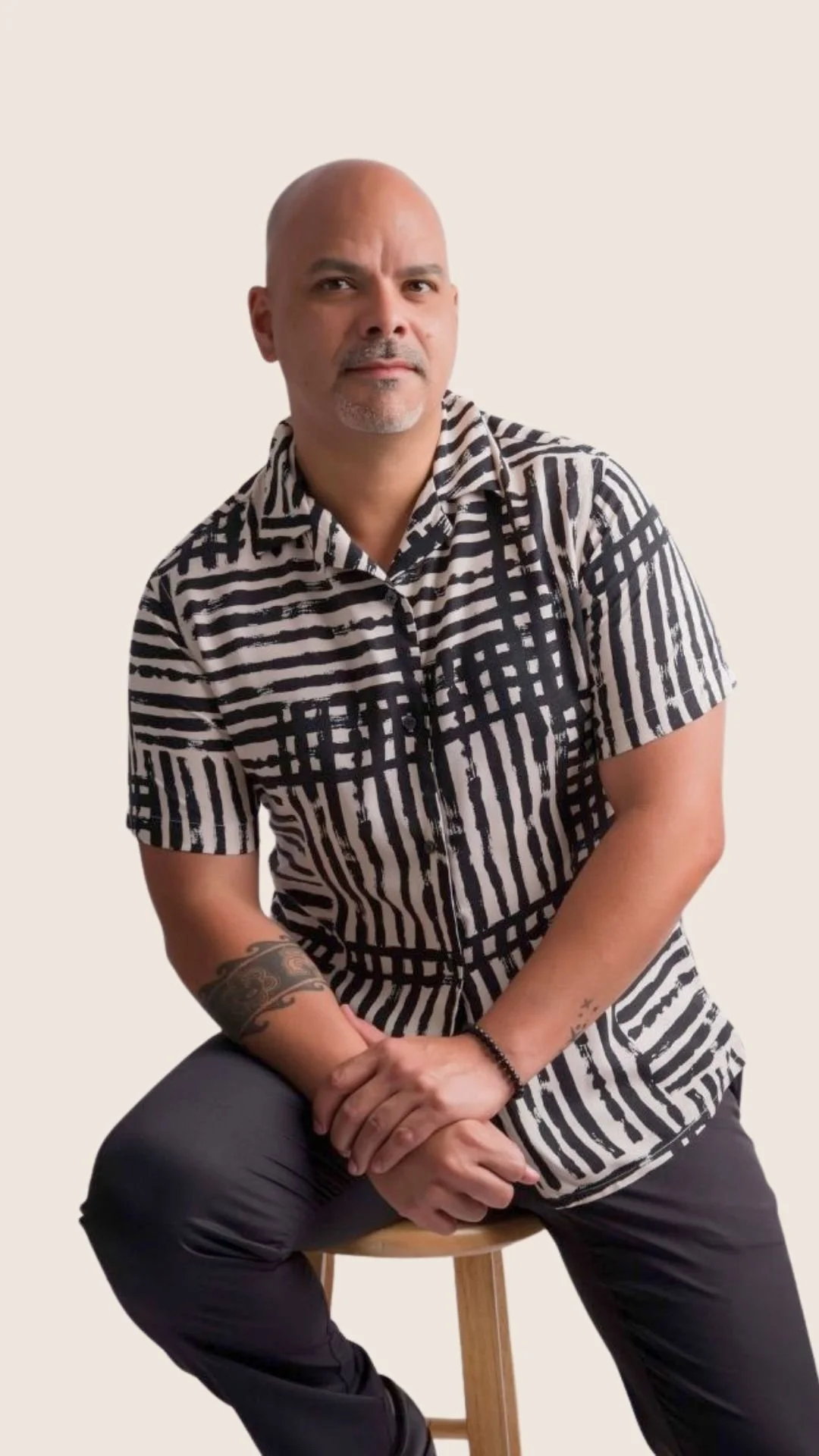 A bald man with a goatee wearing a black and white striped short-sleeve shirt, seated on a wooden stool with hands clasped.