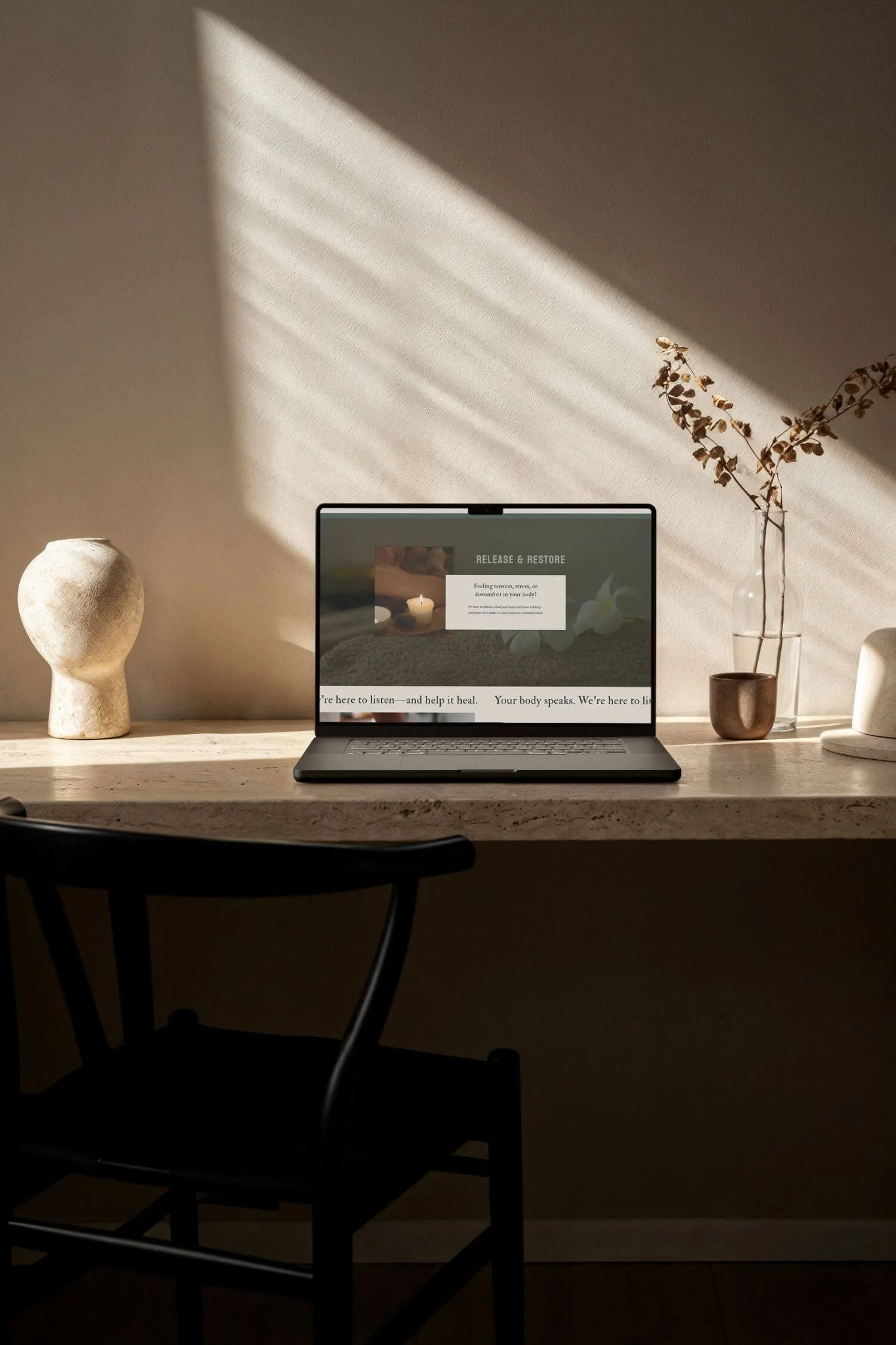 A minimalist desk with a laptop showing the Serene website for massage therapists', a ceramic head sculpture, a glass vase with dried branches, and a small brown pot on a beige surface with lighting casting shadows on the wall.