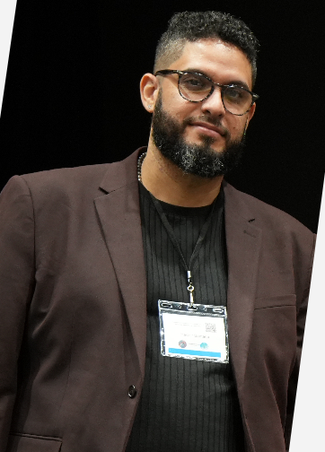 A man with glasses and a beard wearing a dark blazer and black shirt, standing against a dark background.
