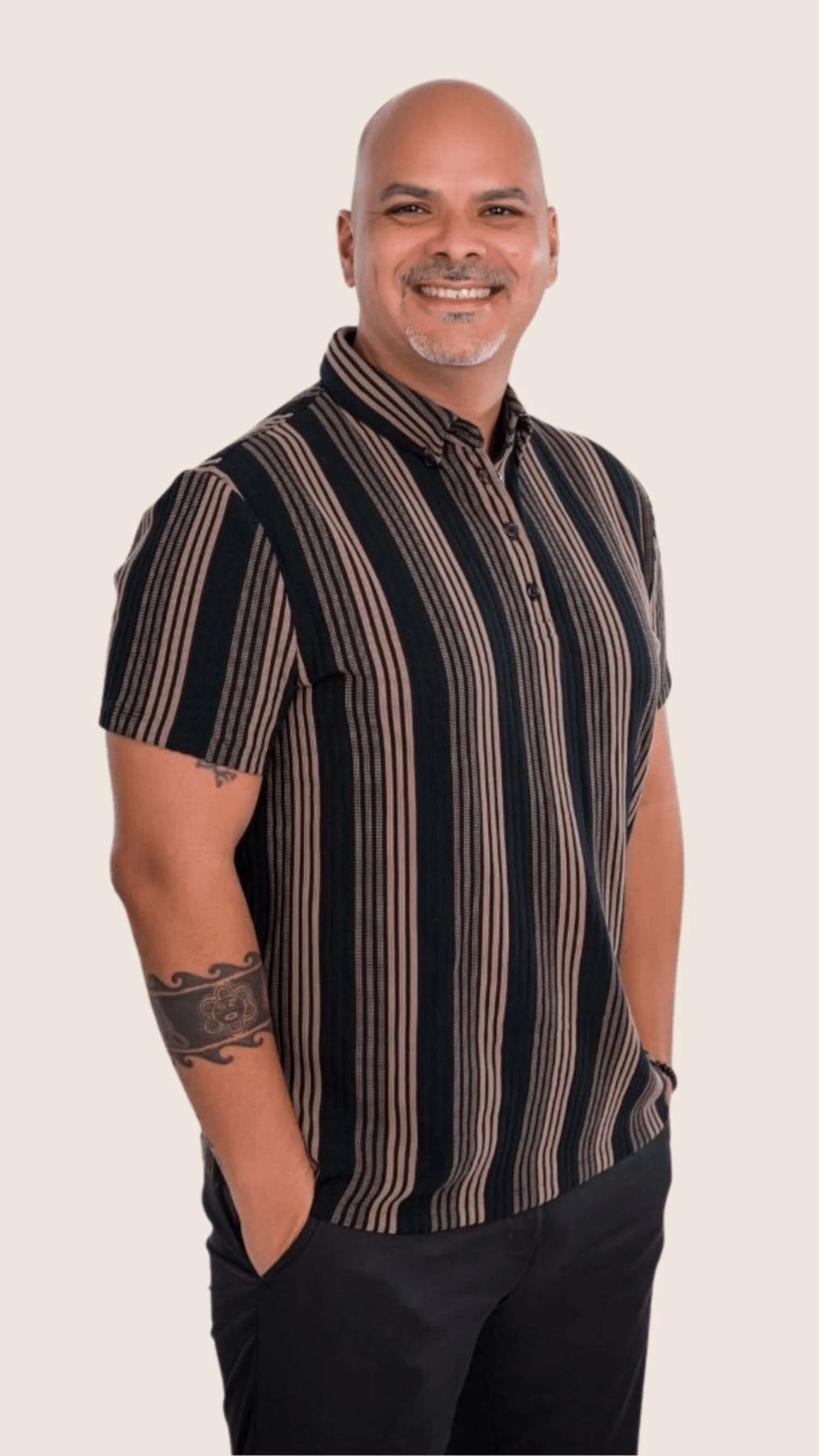 Angel Comas, wearing a black and brown striped short-sleeve shirt and black pants, with a tattoo on his right forearm, standing against a plain light-colored background.