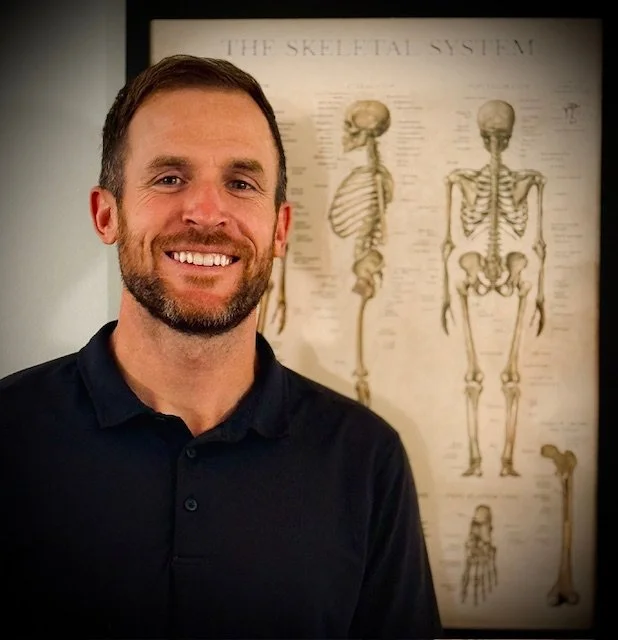A smiling man with a beard in a black shirt standing in front of a poster of the skeletal system.