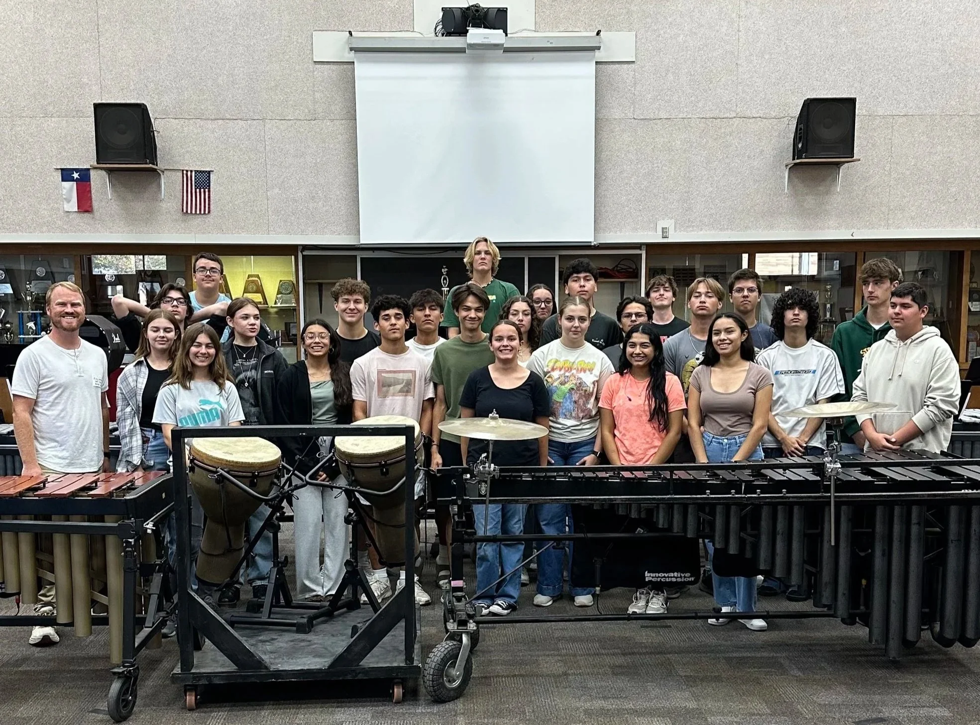 Group of students and two adults posing in a classroom with musical instruments, including drums and xylophones, and a blank projector screen behind them.