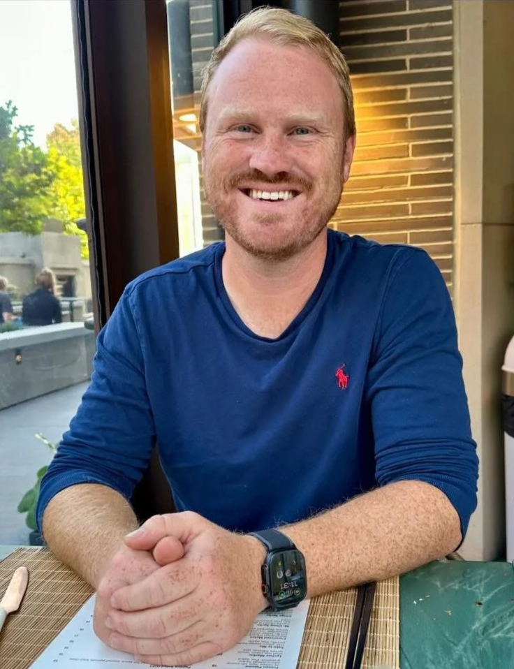 A smiling man with short red hair and beard, wearing a navy blue long-sleeve shirt with a small red logo, sitting at a table in a restaurant or café. He has a black smartwatch on his left wrist and is clasping his hands together on the table. There is a menu and utensils in front of him, with a window and some outdoor views behind him.