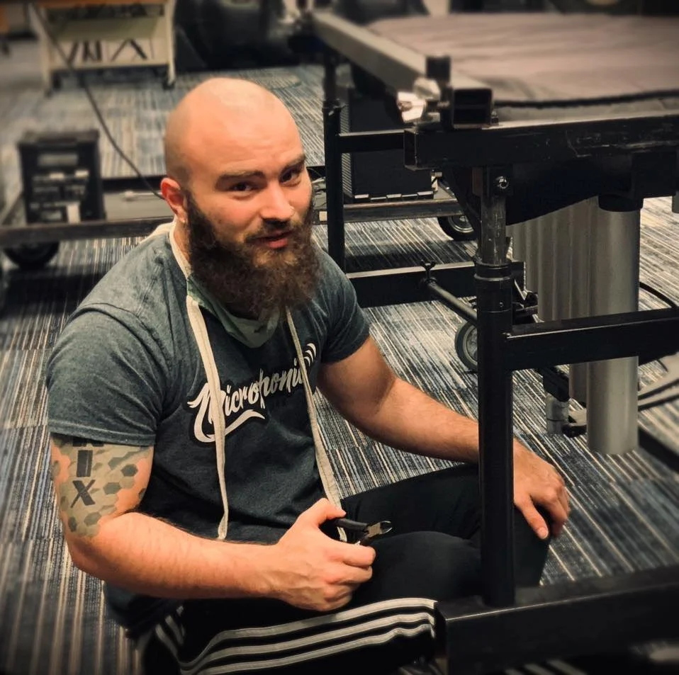 A bald man with a beard, wearing a gray T-shirt and black athletic pants, sitting on the floor next to a black equipment cart, in an indoor setting with carpeted flooring.