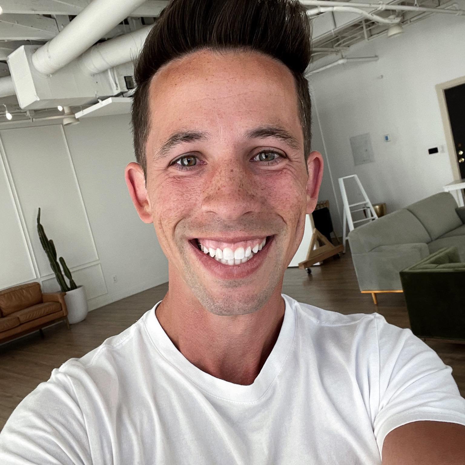 A smiling young man with short brown hair and light skin taking a selfie in a modern, minimalistic living room. The room has white walls, wood flooring, and contemporary furniture including a tan leather couch, a gray couch, and a potted cactus.