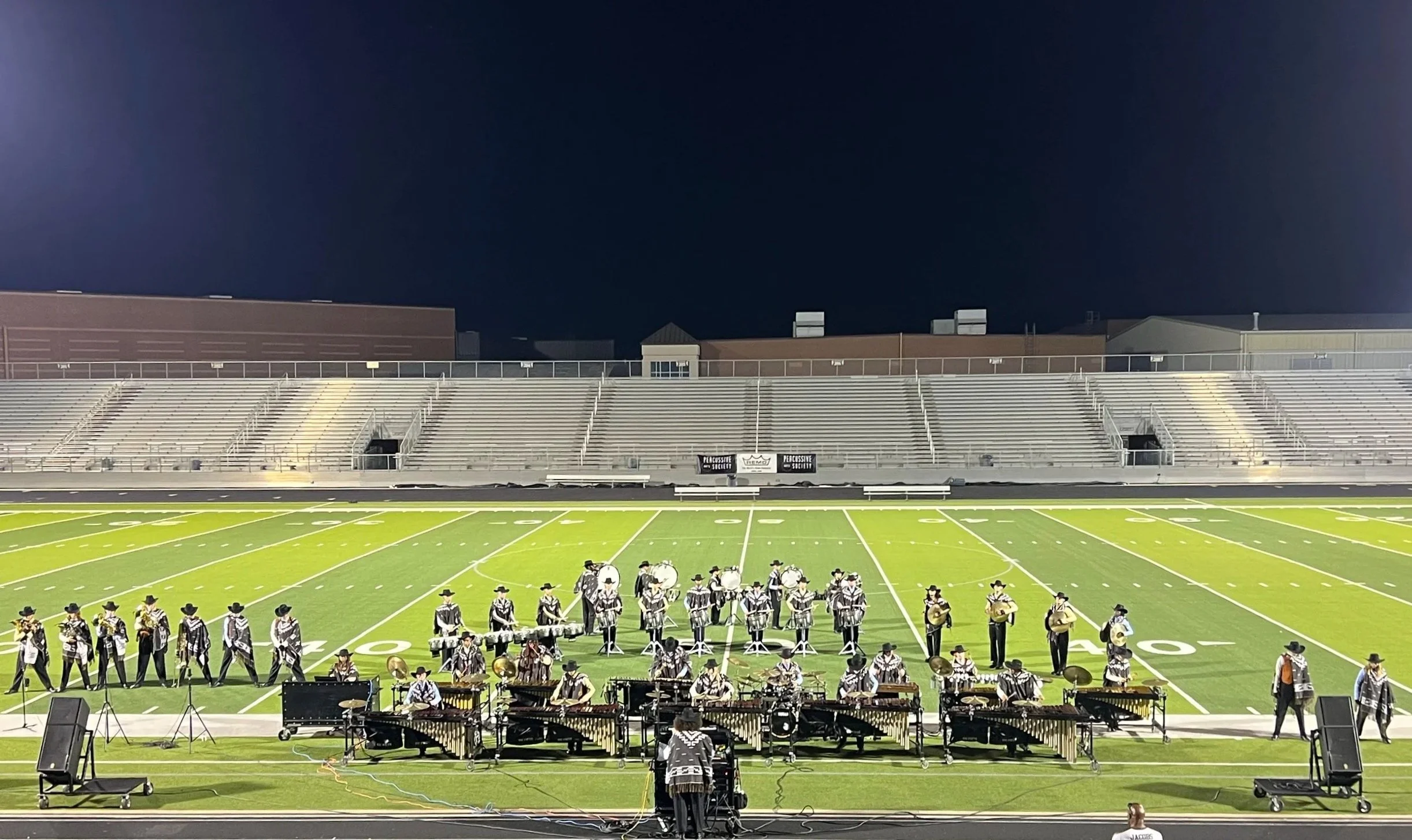A marching band performing on a football field at night, with empty stadium bleachers in the background.