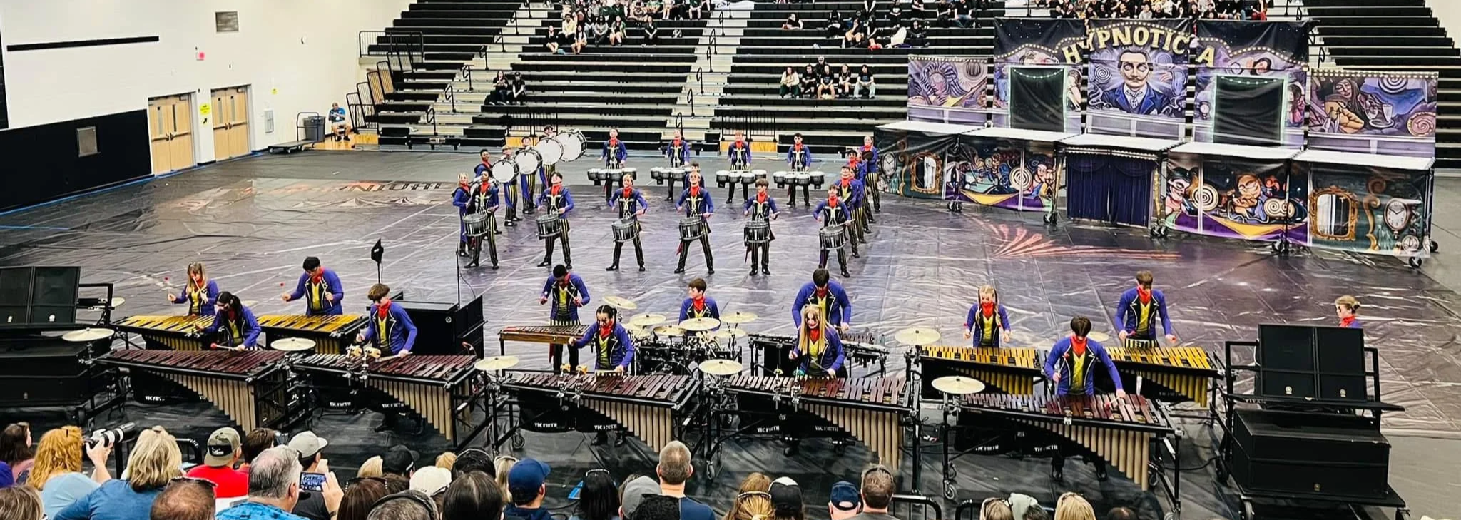 A marching band performance in a gymnasium with audience in the foreground, featuring drummers, percussionists, and other band members on stage and a colorful backdrop.