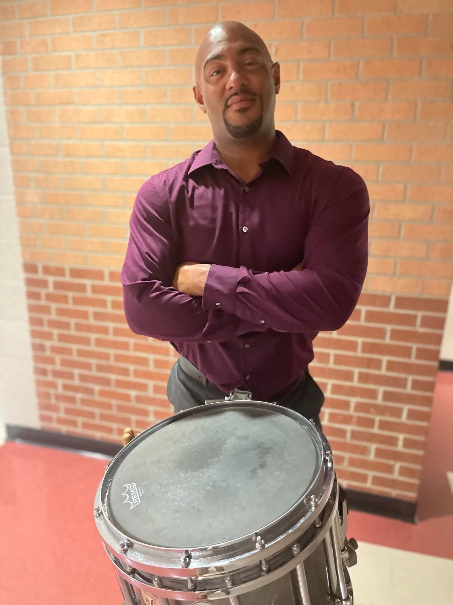 Man with arms crossed standing behind a drum in front of a brick wall.