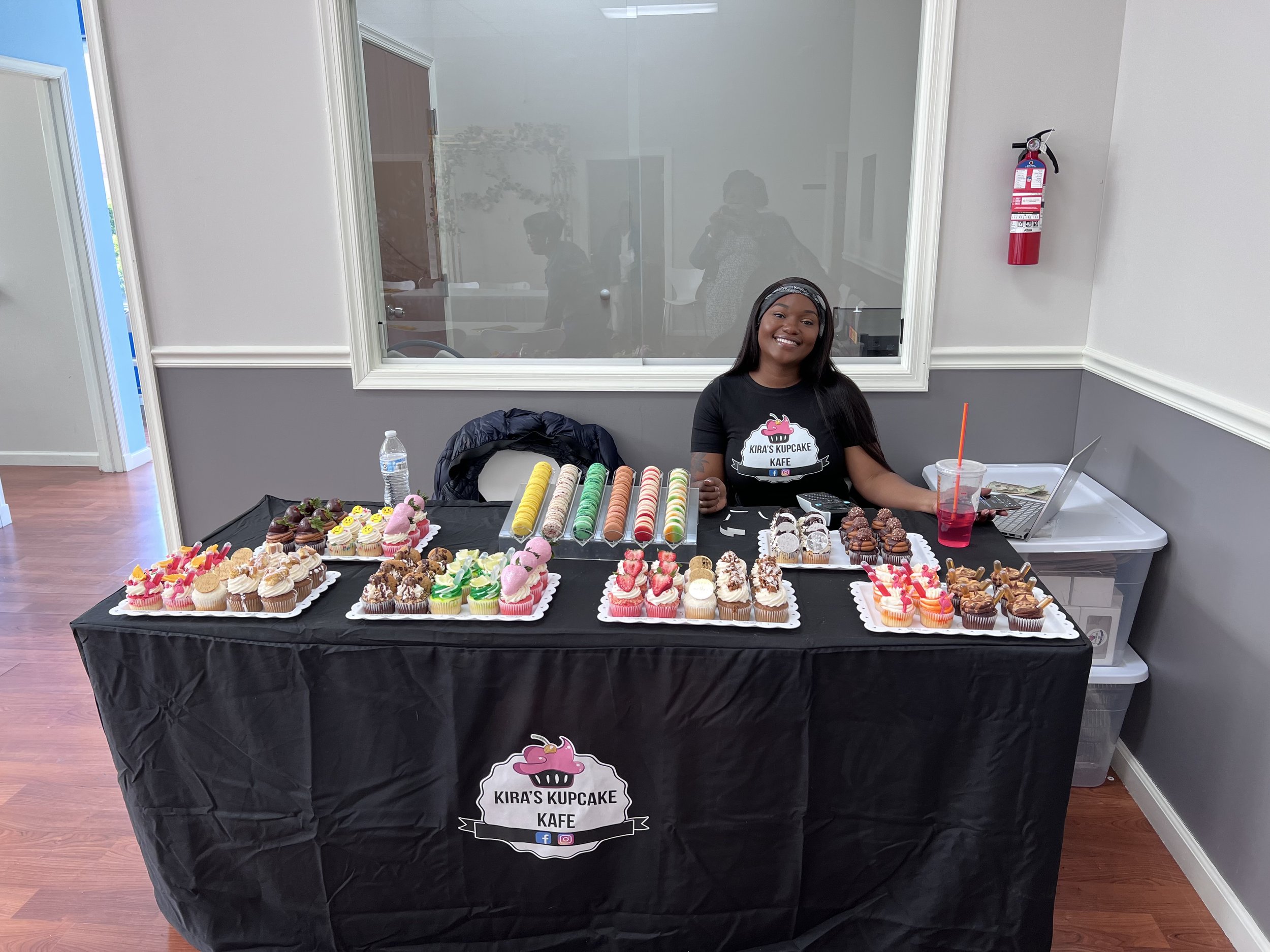 A person sitting behind a table displaying an assortment of cupcakes and macarons. The table is covered with a black cloth featuring the logo "Kira's Kupcake Kafe." Various colorful desserts are arranged on trays in front of the person. The setting a