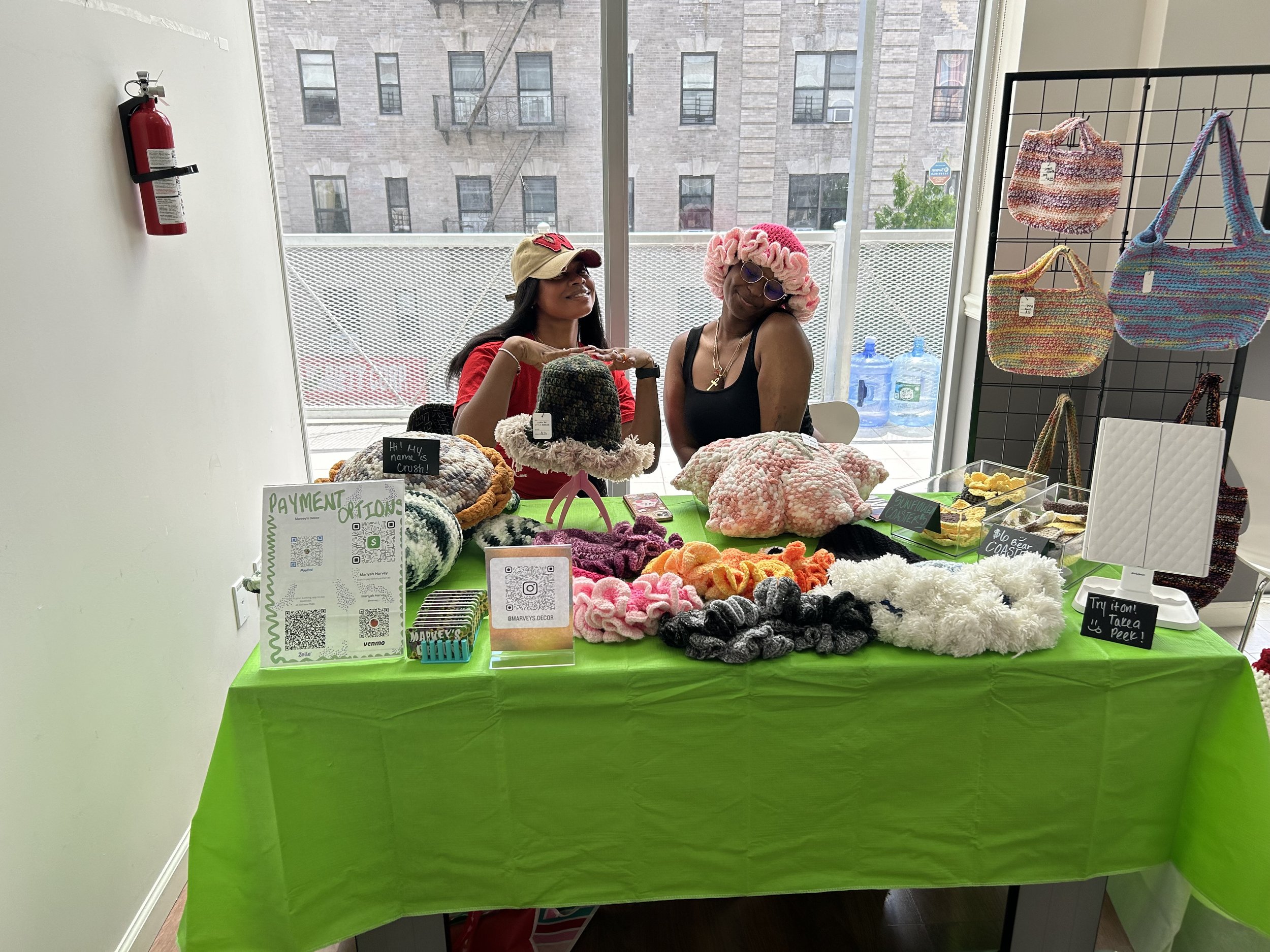 Two people sitting at a table displaying handmade crochet items, including hats and bags. The table is covered with a green cloth and has various crochet products, a payment options sign, and QR codes for digital transactions. Display rack with croch