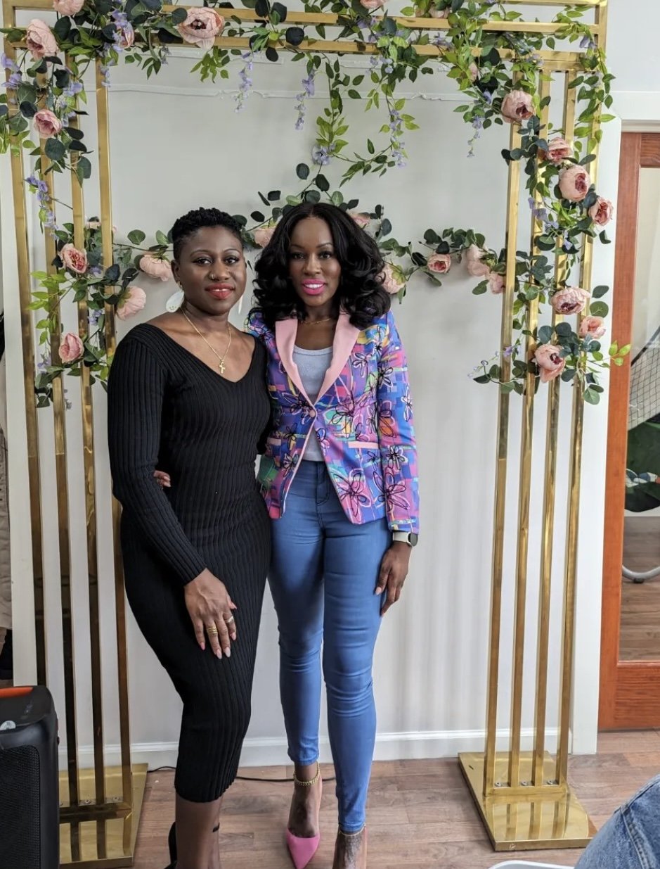 Two women posing in front of a floral archway with gold frames, one wearing a black dress and the other in a colorful blazer and jeans, both smiling.