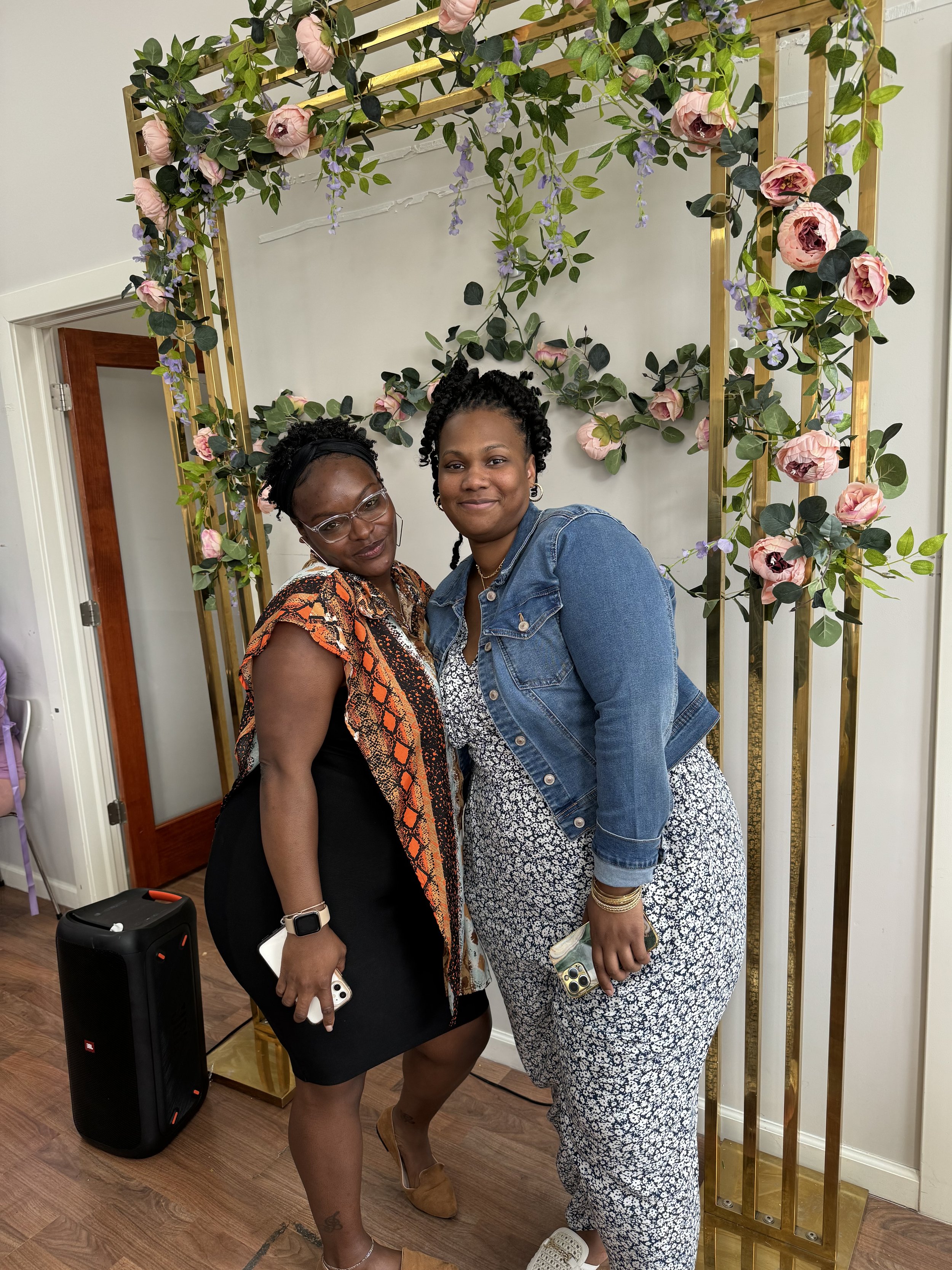 Two women posing in front of a floral arch with pink and green decorations in an indoor setting.