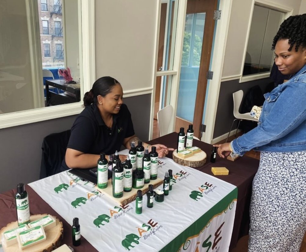 Two people at a table displaying bottles of personal care products with labels, covered by a tablecloth reading "ASE" in a room with mirrored walls.