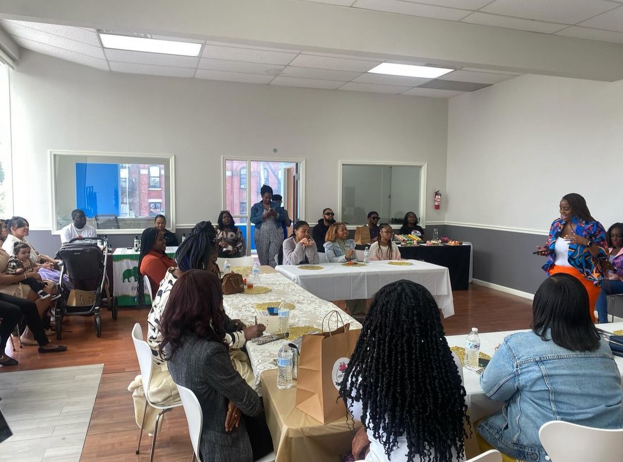 A group of people seated around tables in a bright room, attentively listening to a speaker. The tables are covered with white and gold-colored cloths, and gift bags and bottles of water are visible. The speaker stands on the right, gesturing while o