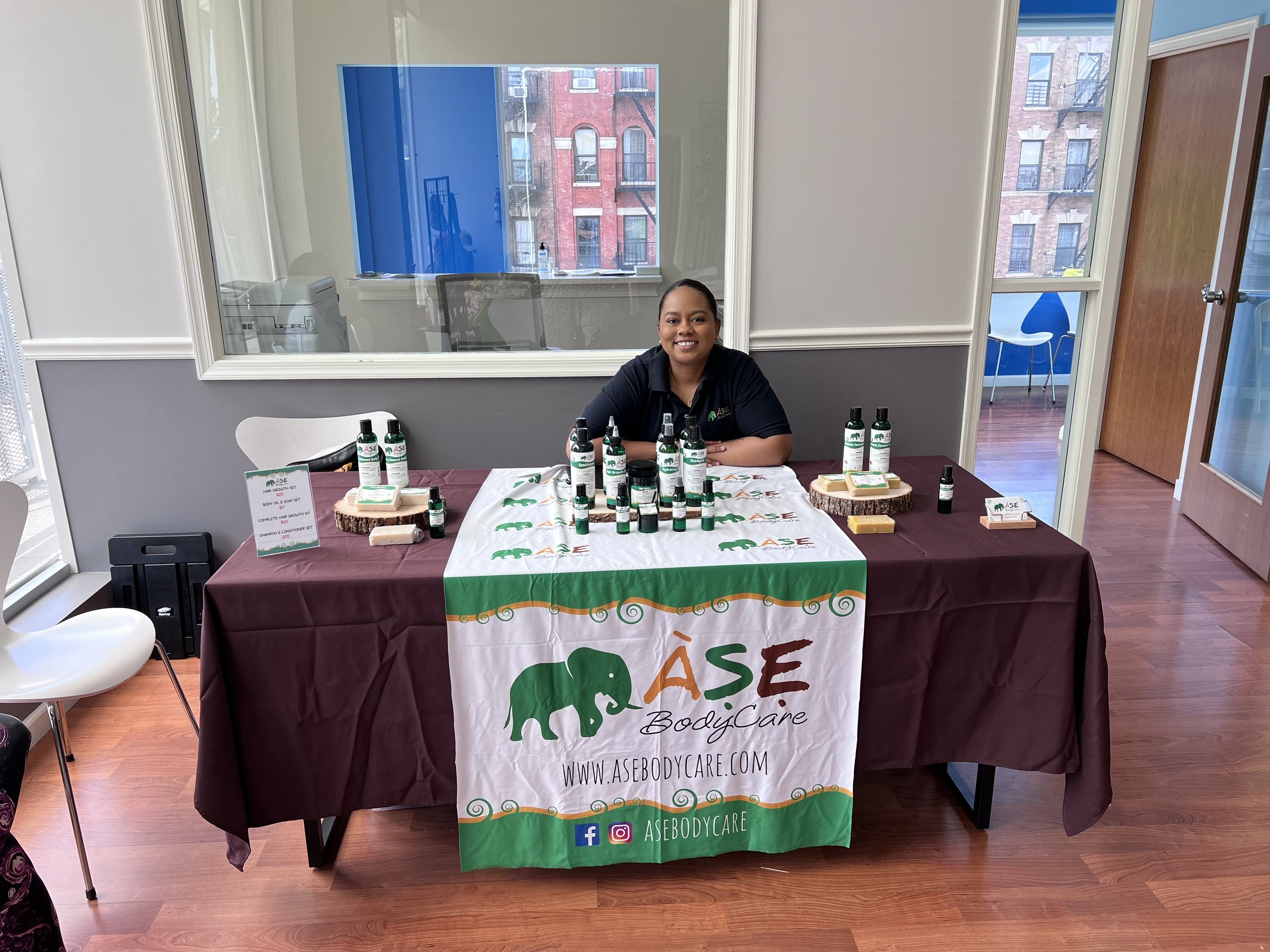 A person sitting behind a booth displaying body care products. The table is covered with a cloth featuring the logo "Ase BodyCare" and several bottles and soaps are arranged neatly. There's a sign and social media information visible on the table. Th