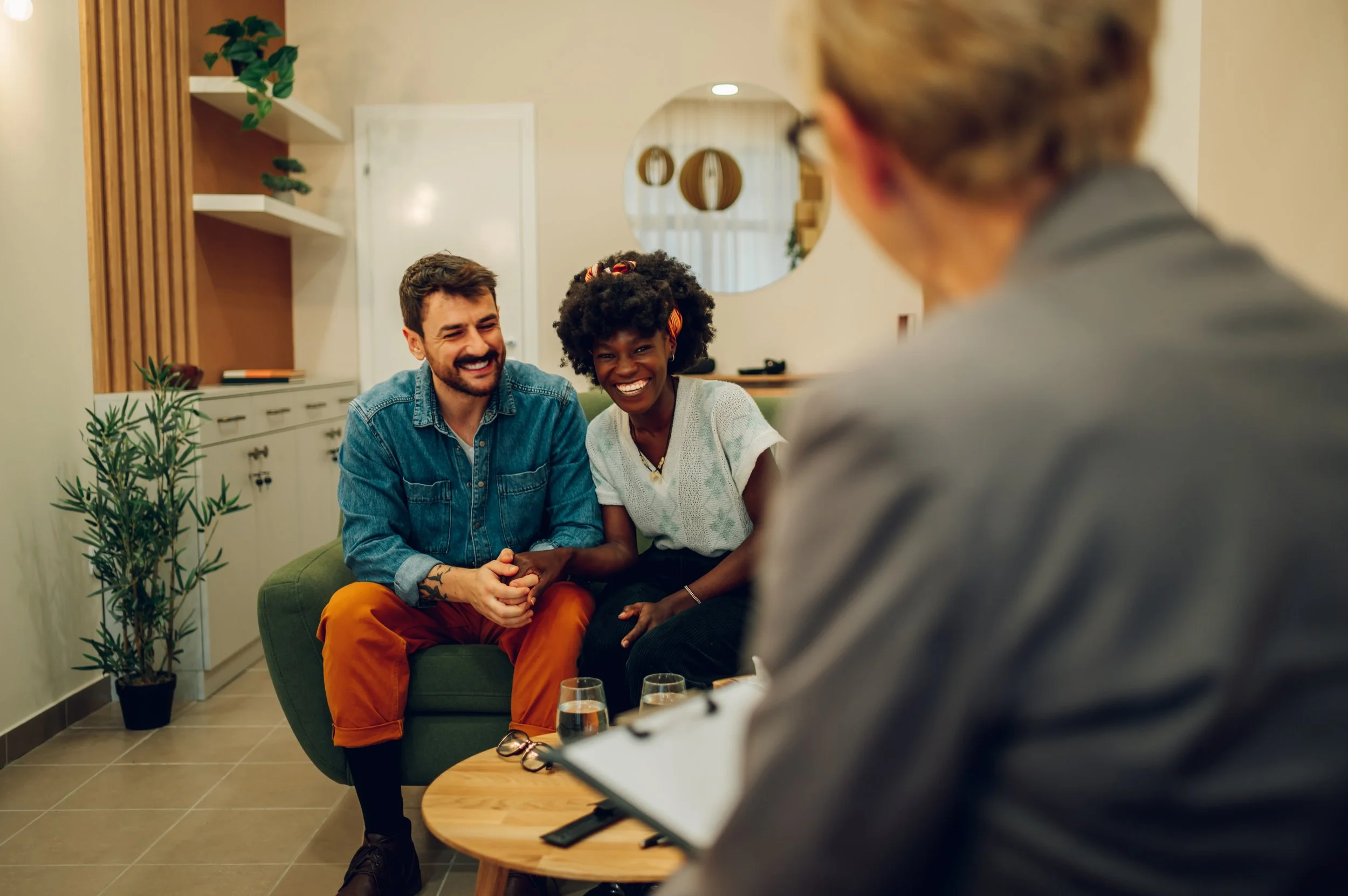 To personer som smiler og holder hender mens de snakker med en annen person som sitter foran dem, i et rom med planter og speil.