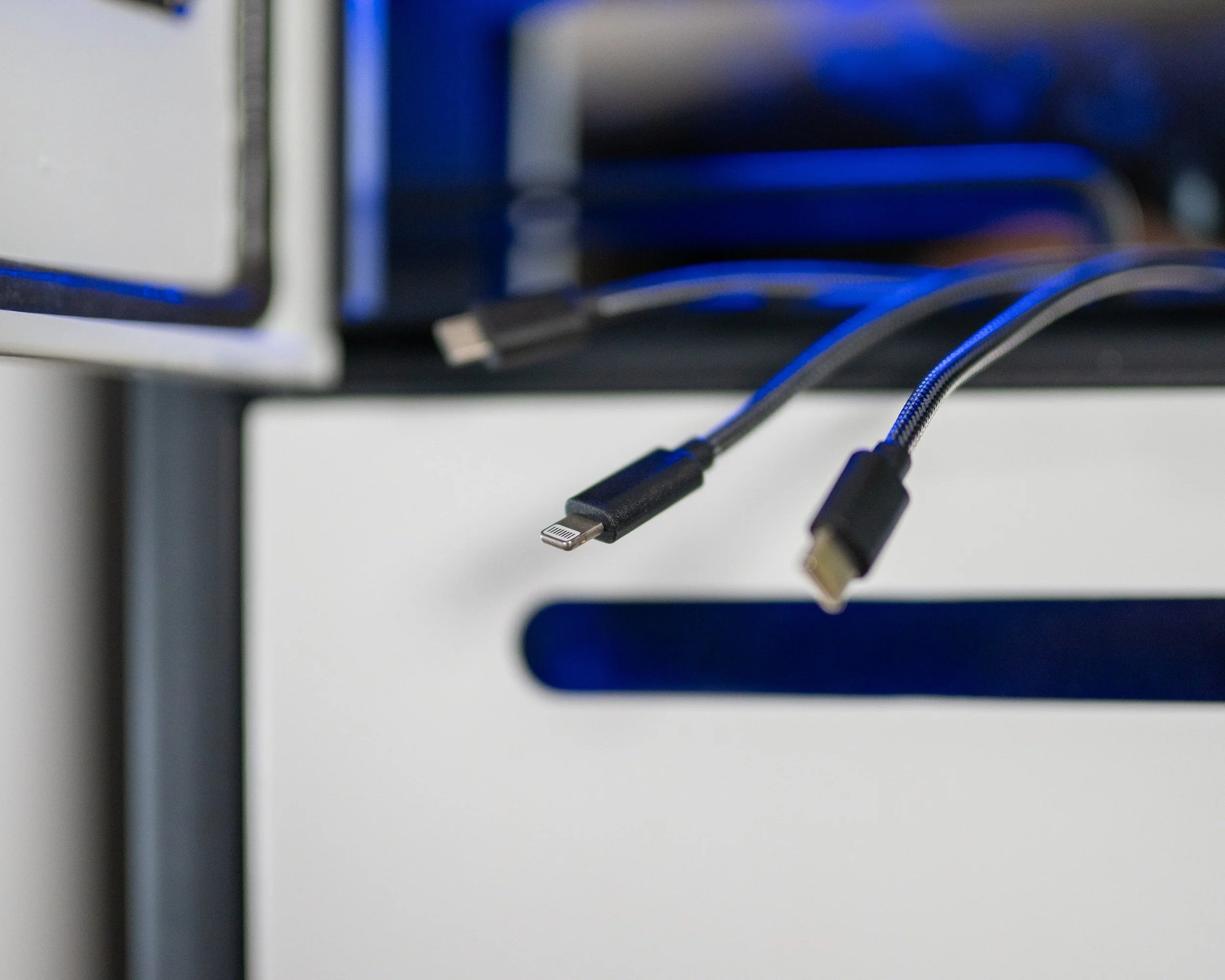 Close-up of two black charging cables with Lightning and USB-C connectors resting on a black surface, with a blurred computer monitor in the background.