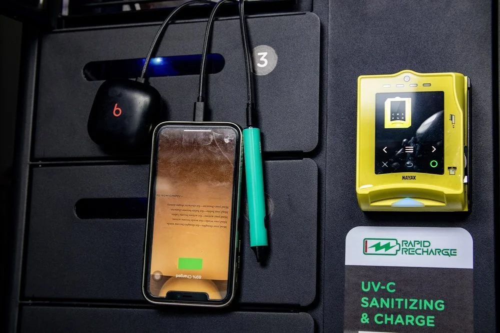 A smartphone charging wirelessly on a black surface, surrounded by a pair of black wireless earbuds in a case, a green pen, a yellow portable device, a warning label about UV-C sanitizing and charging, and a black rectangular object with a blue indic