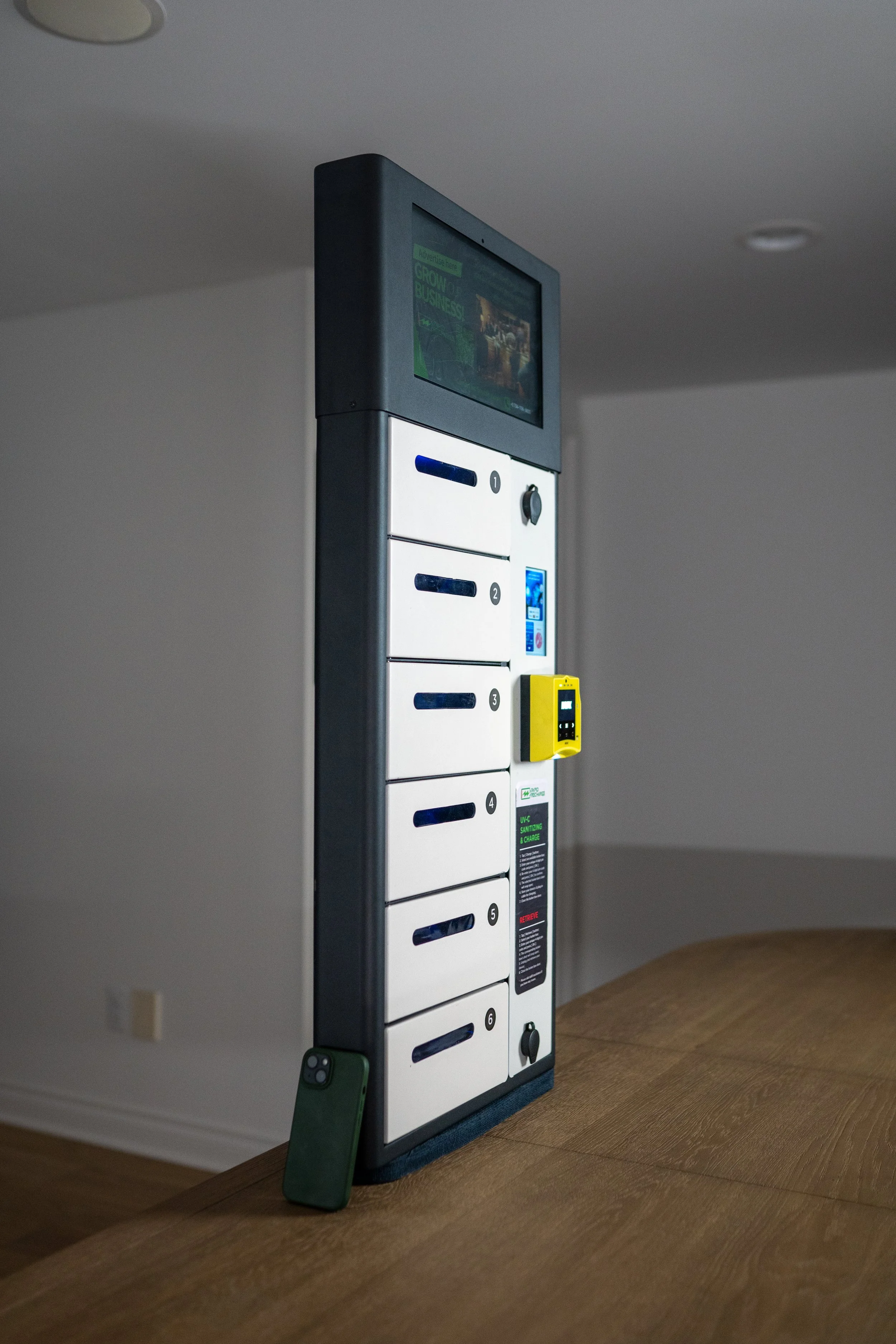 A set of six personal lockers with digital locks, a yellow package box, a display screen, and a smartphone leaning against the locker on a wooden table inside a room.