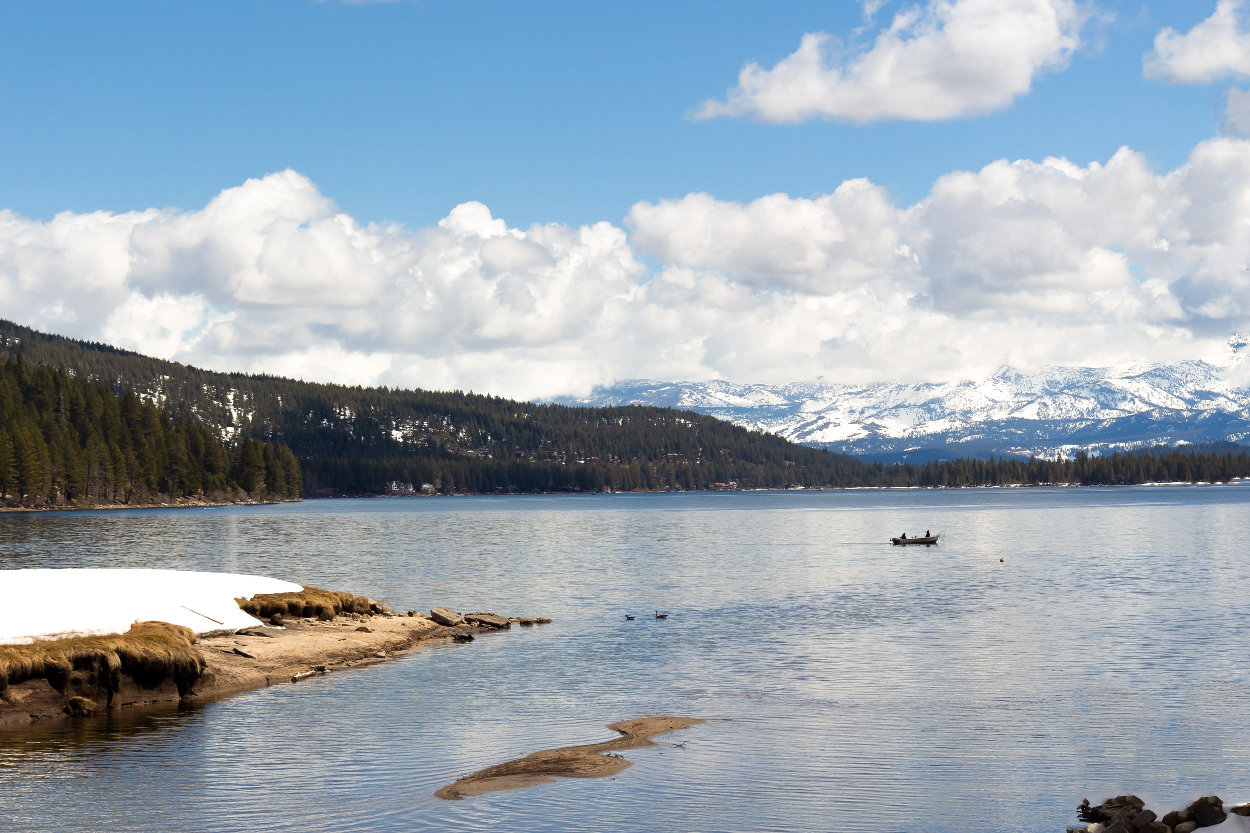 A scenic lakeside view with a small boat on the water, snow-capped mountains in the background, a forested shoreline, and partly cloudy sky.