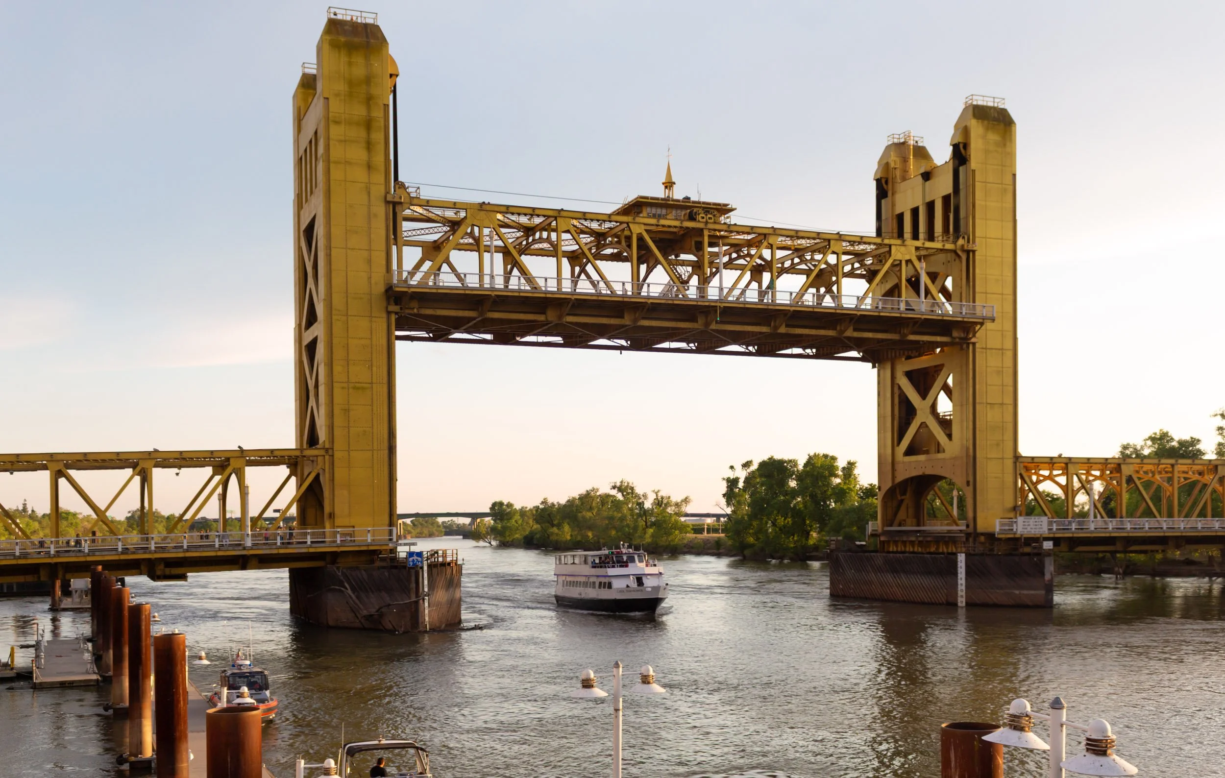 A yellow drawbridge over a river, with a boat passing underneath during sunset, and trees in the background.
