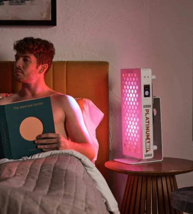 A man with curly hair sitting in bed, reading a book with a dark cover and a large beige circle. He is shirtless and has a pink glow from a nearby pink-colored device on a wooden nightstand. The device has circular holes and the word 'PLATINUM' on the packaging leaning against it.