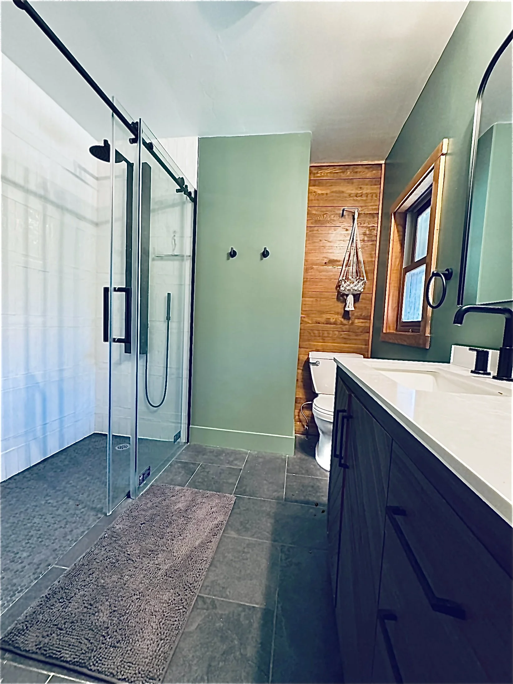 Modern bathroom with tiled floor, glass shower enclosure, and green walls. Features a vanity with a white countertop, black fixtures, and a toilet. A brown rug is on the floor.