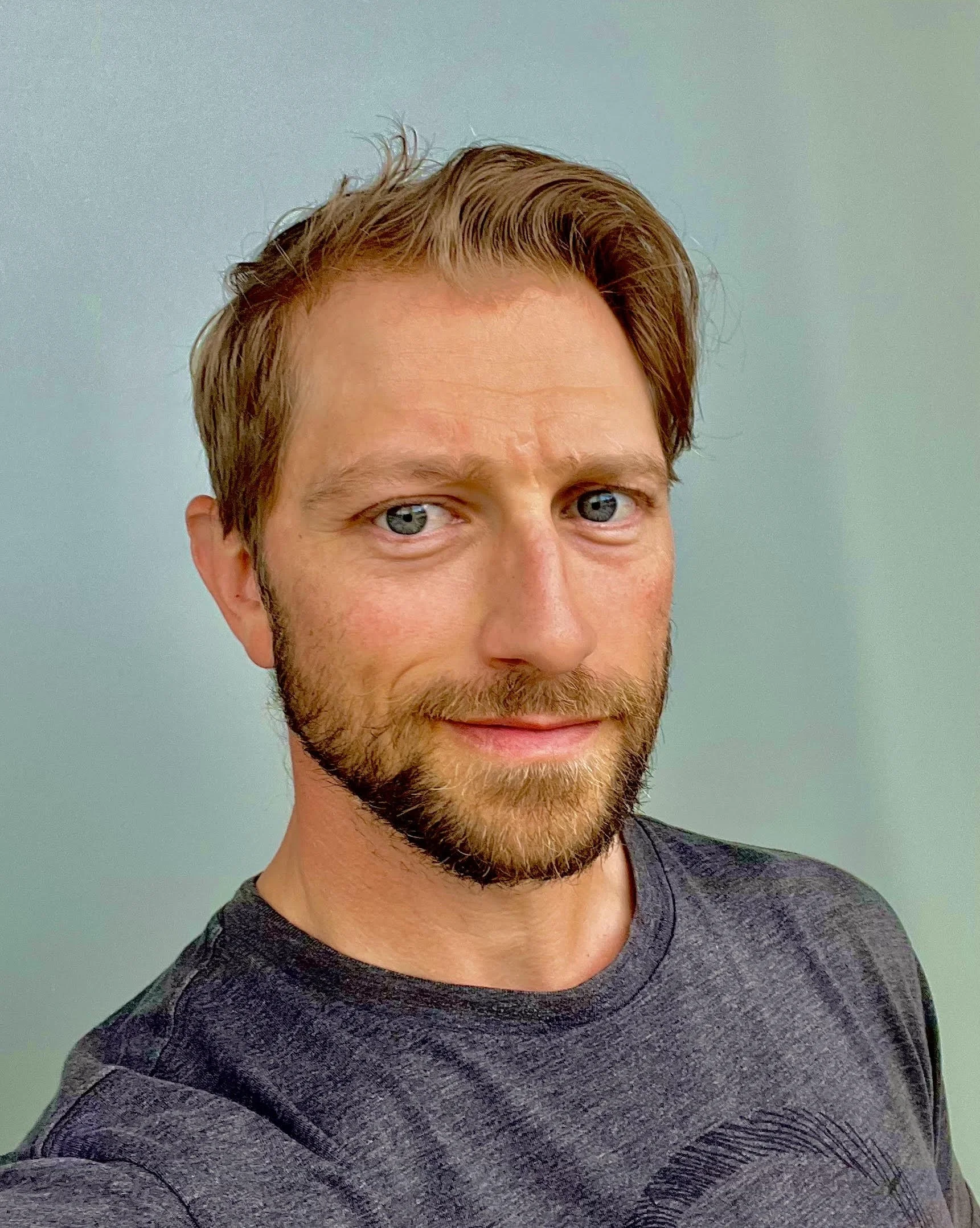 Portrait of a man with short hair and beard, wearing a dark shirt, standing indoors.
