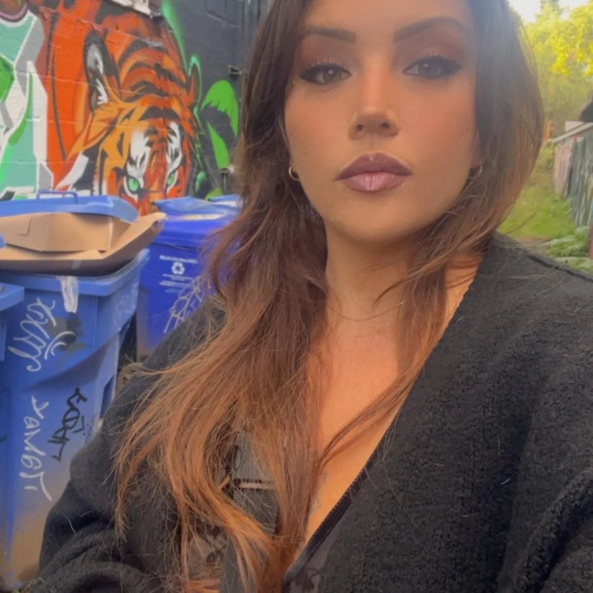 A woman with long, wavy brown hair takes a selfie outdoors. Behind her is a colorful graffiti mural of a tiger with green eyes on a black wall, and several blue recycling bins.