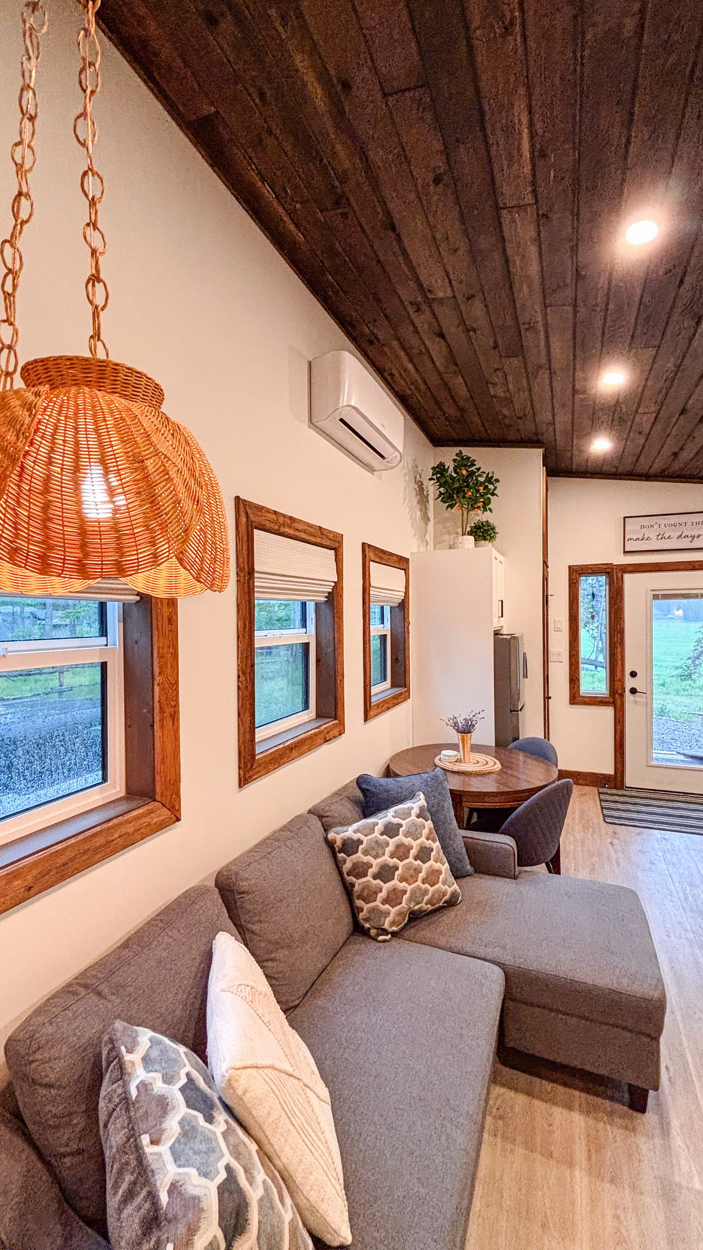Cozy living room with a gray sectional sofa decorated with patterned and plain pillows, a wooden round dining table with chairs, three framed windows with blinds, a wicker hanging lamp, a white cabinet with plants, and a wooden ceiling with recessed lights.