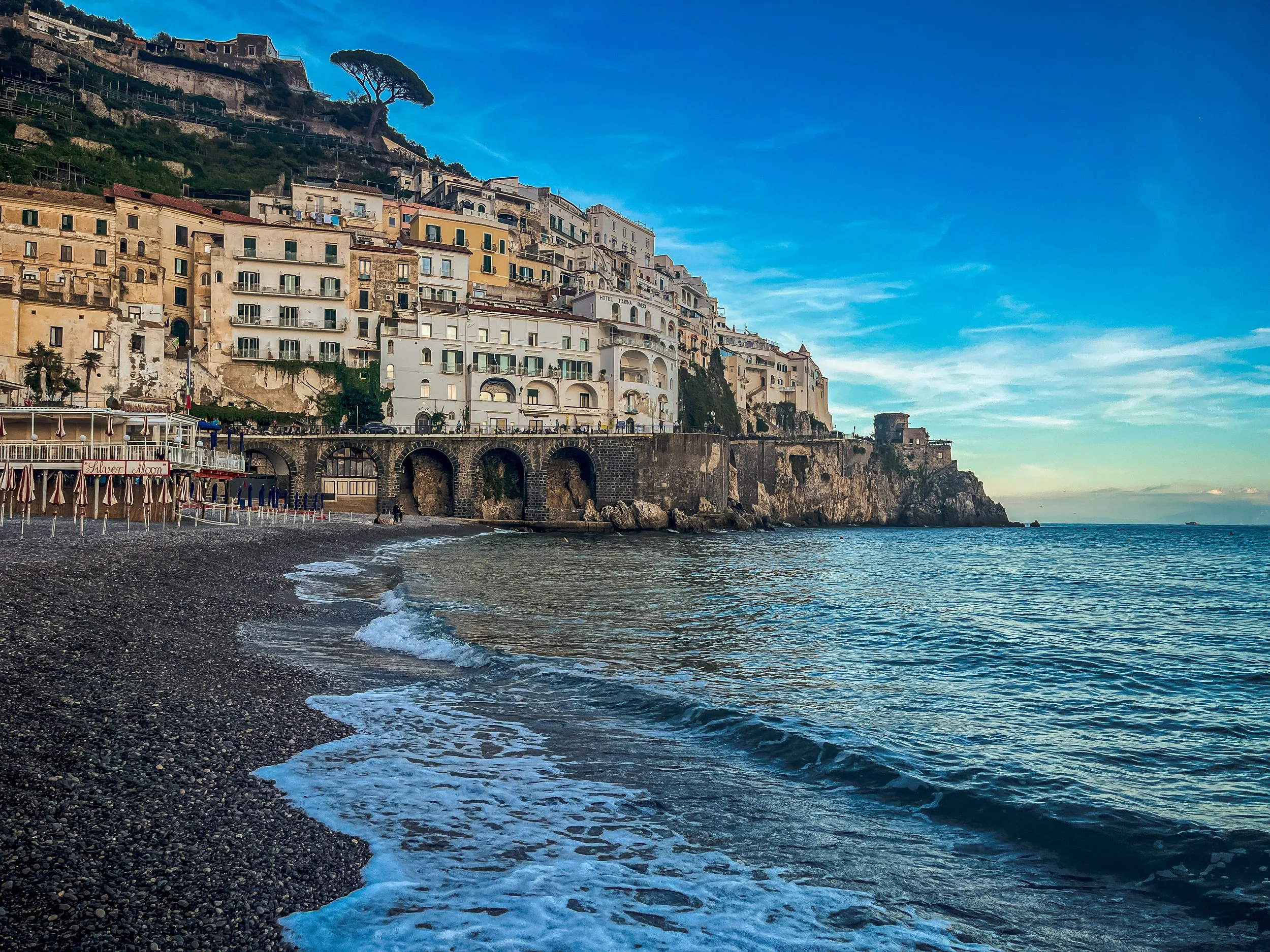 Colorful buildings and houses are built on a steep hillside beside the ocean, with a beach and pebbles in the foreground. The sky is clear with some clouds.