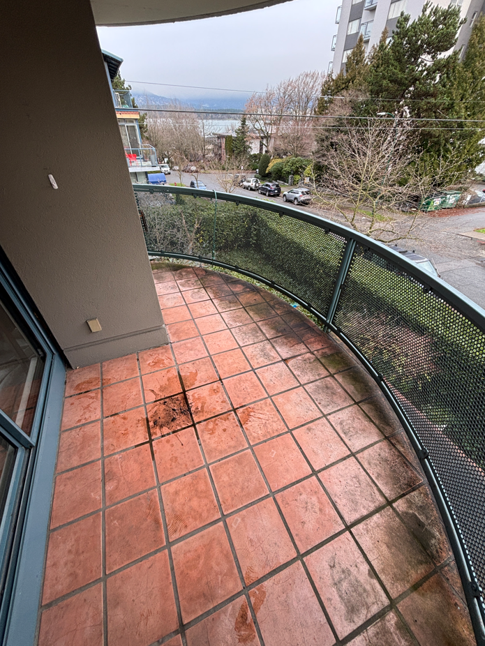 A small, partially damp balcony with concrete flooring, scattered leaves, and a glass railing overlooks a rainy, overcast street lined with trees.