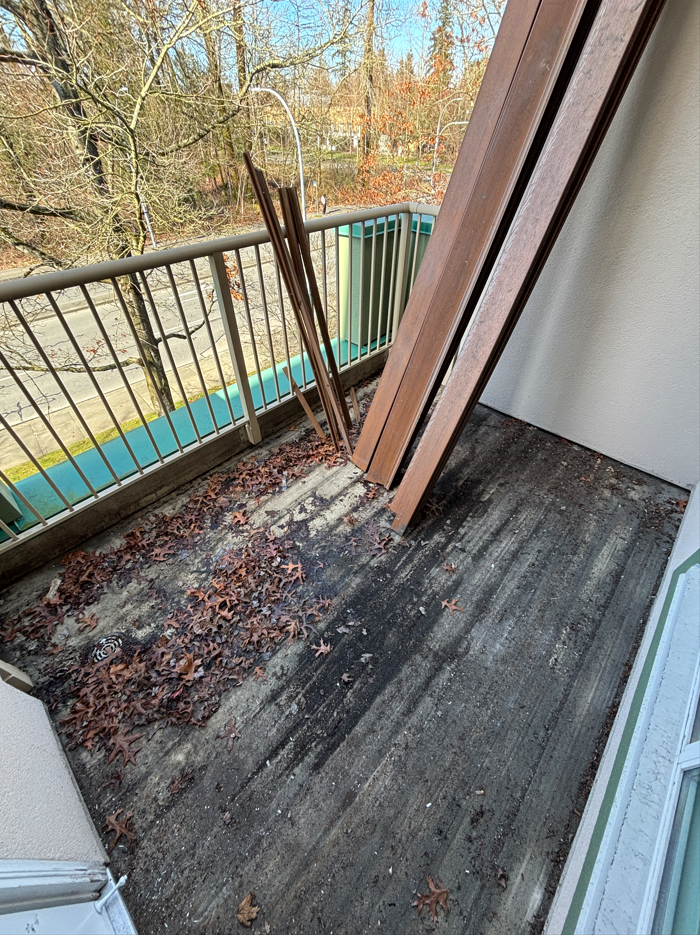 Small apartment balcony with dirty concrete floor, fallen leaves, and four long wooden planks leaning against the railing, overlooking a wooded area.