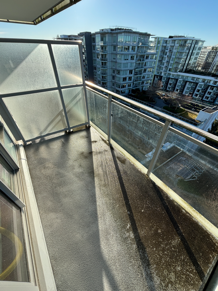A small, partially damp balcony with concrete flooring, scattered leaves, and a glass railing overlooks a rainy, overcast street lined with trees.