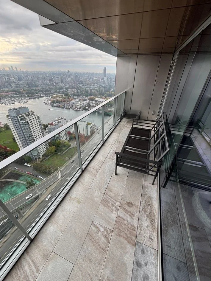 A balcony with three black chairs overlooking a cityscape, with water, boats, and numerous buildings under a cloudy sky.