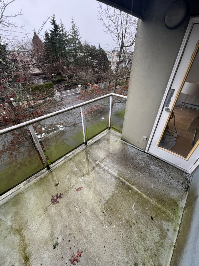 A small, partially damp balcony with concrete flooring, scattered leaves, and a glass railing overlooks a rainy, overcast street lined with trees.