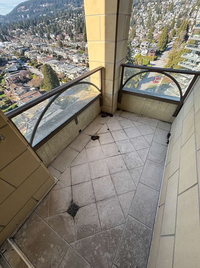 Empty small balcony with beige tiled floor and glass railing, overlooking a city with residential and commercial buildings, surrounded by trees and hills.