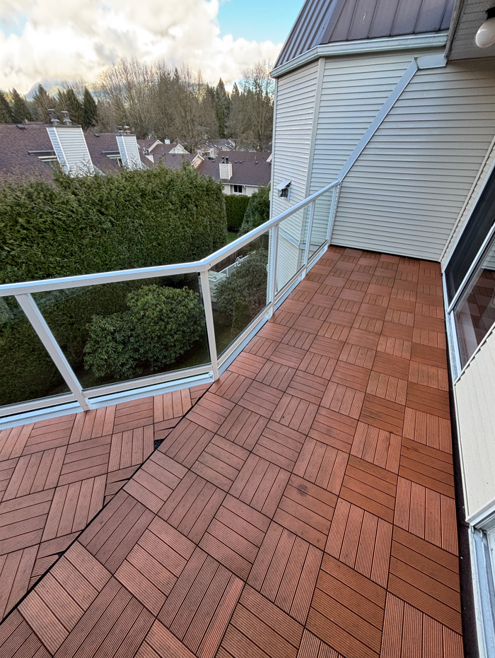 A small balcony with wooden tile flooring, white metal and glass railing, attached to a house with white siding. Houses and trees are visible in the background under a partly cloudy sky.