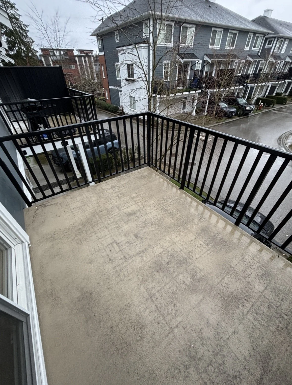 Empty balcony with black metal railings overlooking a wet street and neighboring residential buildings on a cloudy day.