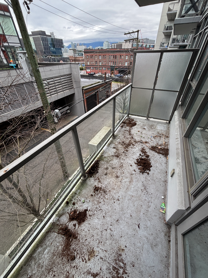 Empty balcony with dirt and debris on the concrete floor, surrounded by a glass and metal railing, overlooking a city street with buildings and power lines under a cloudy sky.