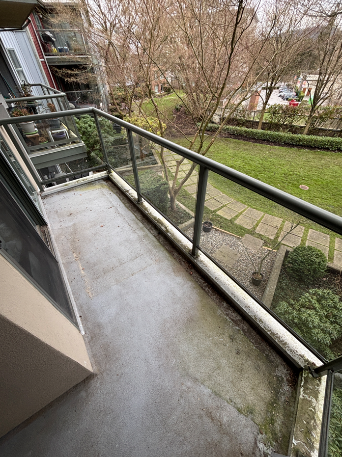 View of a small apartment balcony with a glass railing overlooking a grassy yard with trees and a stone walkway.