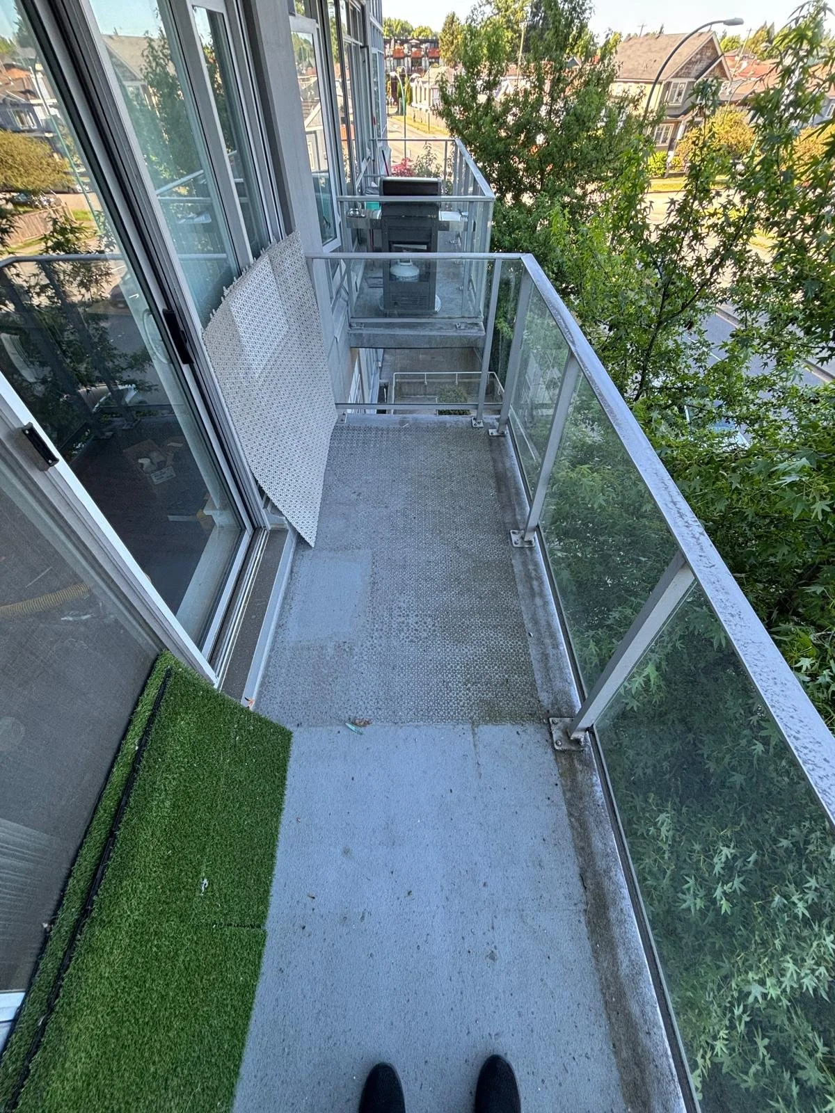 Empty apartment balcony with glass railing, a small patch of artificial grass, and a view of trees and neighboring houses.