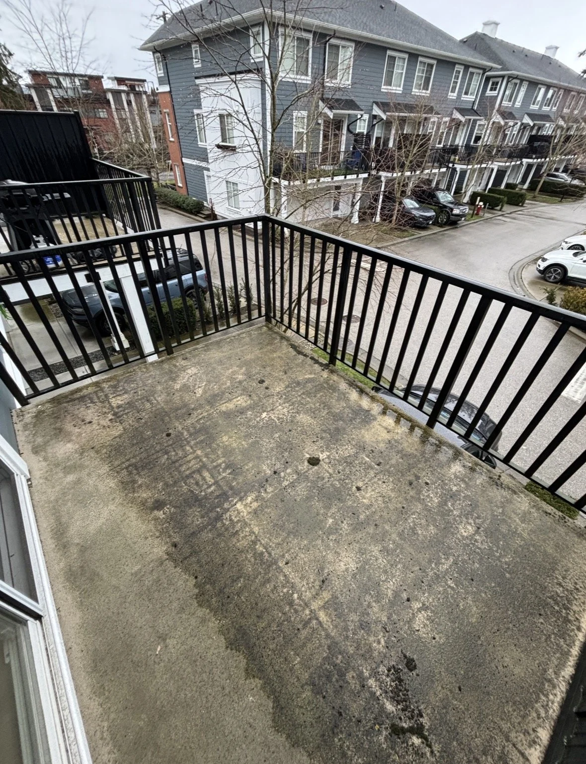 A small, partially damp balcony with concrete flooring, scattered leaves, and a glass railing overlooks a rainy, overcast street lined with trees.