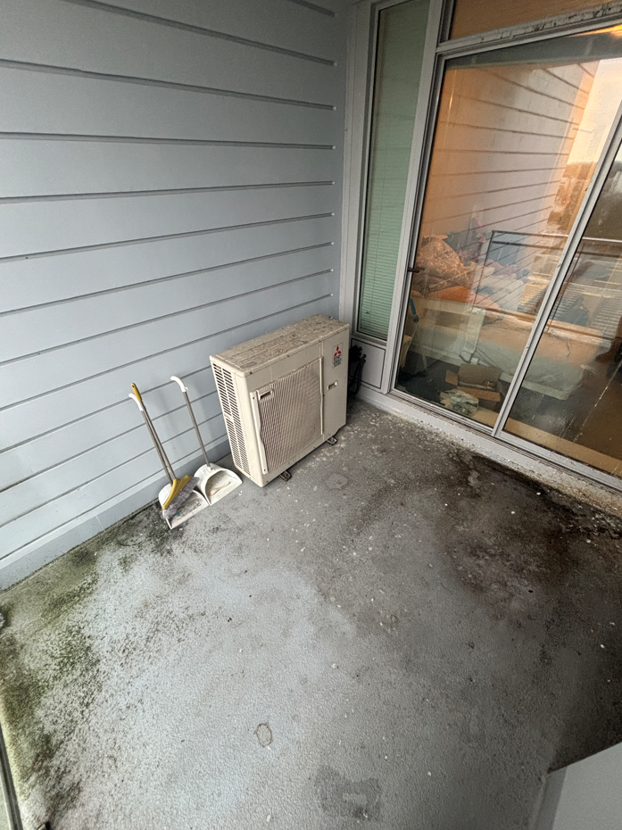 A small, partially damp balcony with concrete flooring, scattered leaves, and a glass railing overlooks a rainy, overcast street lined with trees.