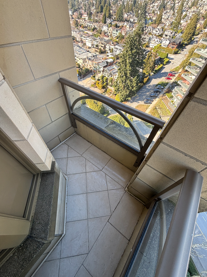 Small balcony with tiled floor, metal railing, and glass wind guard, overlooking a cityscape with houses, trees, and cars.