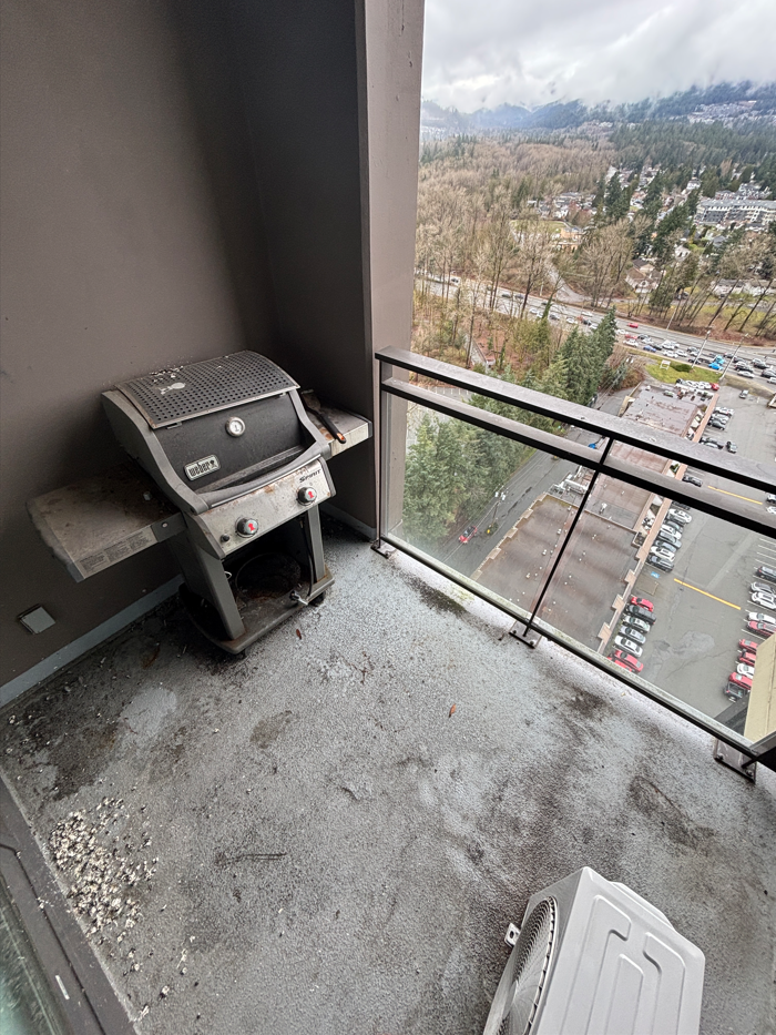 Balcony with a used barbecue grill and a white air conditioning unit, overlooking a parking lot and trees with mountains in the background.