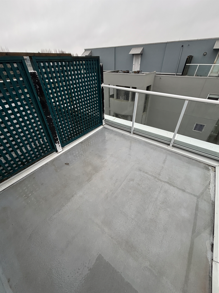 A small, empty terrace with wet, gray flooring and white railings overlooks a neighboring building. A dark green lattice fence adds contrast on the left. Cloudy gray sky suggests recent rain.